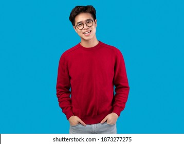 Casual Teenager. Portrait Of Smiling Asian Guy Wearing Glasses And Red Sweater, Standing With Hands In Pockets Isolated Over Blue Studio Background. Cheerful Male Model Posing And Looking At Camera