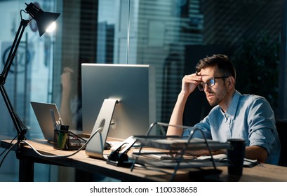 Casual tired office worker sitting at desk using computer and doing overtime project in lamplight. 
