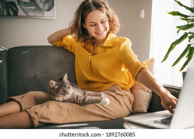 Casual woman in yellow shirt working on laptop with her cat on sofa, sitting together in room with window ang green plant