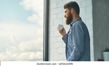 Casual young bearded man drinking coffee at work Coffee really gives more energy. Bearded man enjoy coffee in morning looking at the window Freelance people home work lifestyle with copy space concept