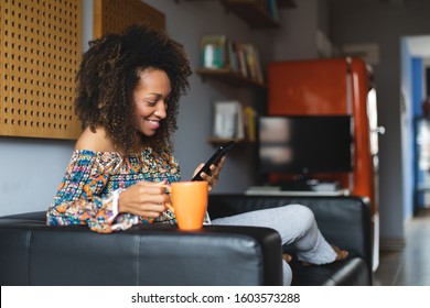 Casual young black woman relaxing at home texting on smartphone and drinking coffeee. Home leisure and tranquility concept. Afro hairstyle model.