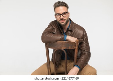 casual young guy in brown leather jacet holding arm on chair and posing in front of grey background 