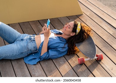 Casual young hipster girl messaging in smartphone using wireless 4g internet connection outdoors lying relaxed in longboard at summer sunset. Trendy dressed stylish female with mobile phone in hands