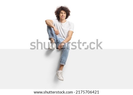 Casual young man with curly hair sitting on a blank panel isolated on white background