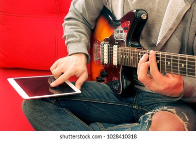 Casual young man in ripped jeans, playing sunburst offset electric guitar. Young musician taking online musical instrument course lesson on wireless internet, tablet gadget. Background, close up. - Powered by Shutterstock