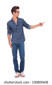 Casual Young Man Standing With A Thumb In His Pocket And Pointing And Looking To His Side With A Smile On His Face. Isolated On A White Background