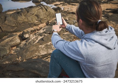 A casual young man uses his smartphone while sitting on a rocky coastline. The serene outdoor setting signifies a blending of technology and nature, highlighting a moment of solitude and reflection. - Powered by Shutterstock