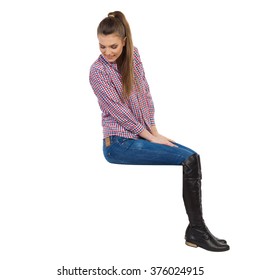 Casual Young Woman In Lumberjack Shirt, Jeans And Black Boots Sitting On A Top And Looking Down. Side View. Full Length Studio Shot Isolated On White.