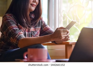 Casual young woman in plaid striped shirt hands holding and using mobile smart phone and work on computer notebook with pink cup of coffee on the wooden table near window in morning day light at home.