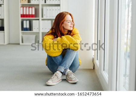 Casual young woman relaxing at the office sitting on the floor in front of a glass door facing outside with closed eyes and a blissful smile