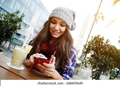 Casual Young Woman Sending Text Message While Sitting At A Sidewalk Cafe In The City, Charming Hipster Girl Enjoying A Fresh Cocktail During Her Break Or Holiday, Female Student Chatting On Cell Phone