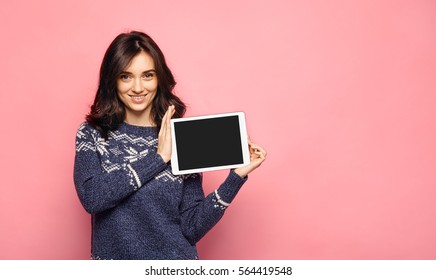 Casual young woman showing a blank tablet screen. Girl in warm winter sweater smiling and holding a tablet with empty black display for your content