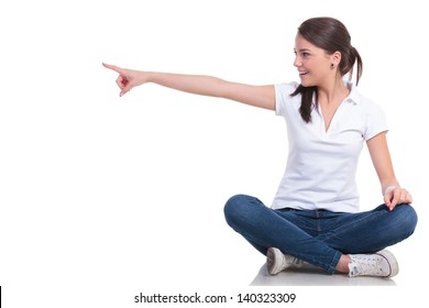 Casual Young Woman Sitting With Legs Crossed Pointing And Looking To Her Side With A Smile. Isolated On White Background