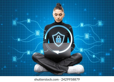 Casual young woman sitting on floor and using laptop with glowing padlock shield hologram on blurry blue background. Safety, web protection and antivirus concept