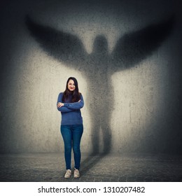 Casual Young Woman Student Full Length Portrait Holding Arms Crossed Confident Looking As Casting A Hero Shadow With Angel Wings On A Dark Room Wall. Inner Strength, Ambition And Leadership Concept.