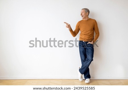 A casually dressed man stands against a blank wall, pointing to something off-camera with a look of interest or emphasis