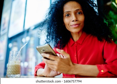 Casually Dressed Mature Black Woman In Red Blouse With Afro Hairstyle Looking Away From Text Messaging Via Smartphone And Smiling