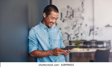 Casually dressed young Asian designer smiling and using a digital tablet while leaning against a gray wall in a modern office