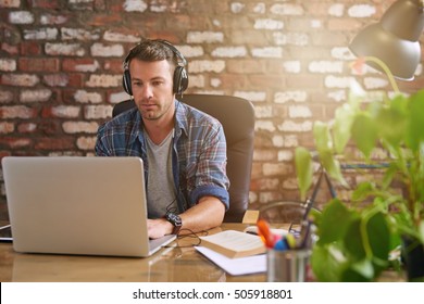 Casually dressed young designer listening to music on headphones while working on a laptop at a desk in an office   - Powered by Shutterstock