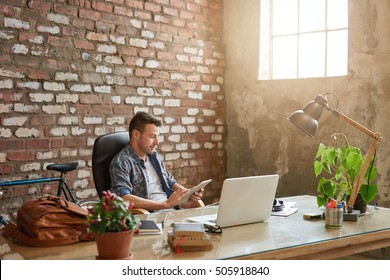 Casually dressed young designer using a tablet while sitting at a desk in a brick walled office   - Powered by Shutterstock