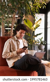 Casually Dressed Young Man Checking Mobile Phone Working On Laptop In Seating Area Of Modern Open Plan Office