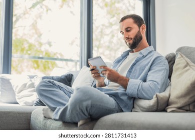 A casually dressed young man sits comfortably on a sofa by a bright window, writing in a small notebook and thoughtfully planning ideas. Warm, relaxed lifestyle scene suggesting creativity and focus at home.