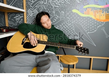 Casually dressed young man strums his guitar in a lively recording studio, lost in music.