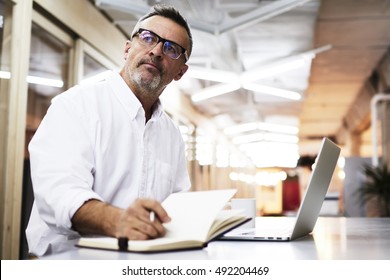 Casually-dressed mature businessman thinking about write news article while working late in an office. Creative editor is working on a new project. Skilled businessman in eye glasses making report - Powered by Shutterstock