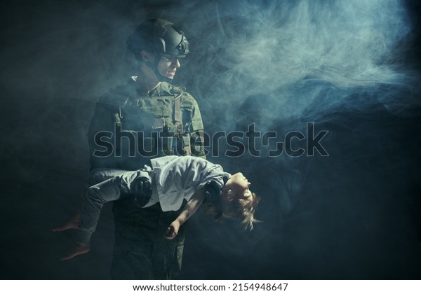 Casualties of war. Portrait of a\
courageous soldier carrying a little girl in his arms injured in\
war zone. Studio portrait on a dark background with smoke. Copy\
space.
