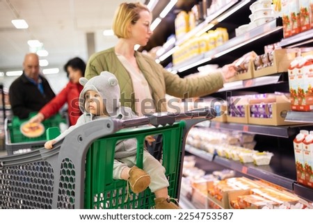 Casualy dressed mother choosing products in department of supermarket grocery store with her infant baby boy child in shopping cart