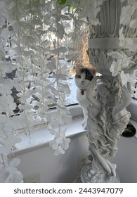 A cat peeking behind an Italian sculpture with a snowy background and white flower backdrop