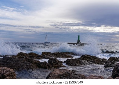 Catamaran In High Seas On The Croatian Mediterranean Coast. The Waves Crash Against The Rocky Coast. In The Background A Green Lighthouse And Dark Clouds. Dramatic Scenery.