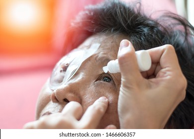 Cataract Treatment After Surgery By Caregiver Concept. Caregiver Use Ophthalmic Solutions Or Eye Drops At Asia Old Woman Patient Eyeball With Protective Shield Over On Other Side.