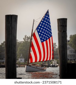 A catboat with an American Flag sail in a harbor