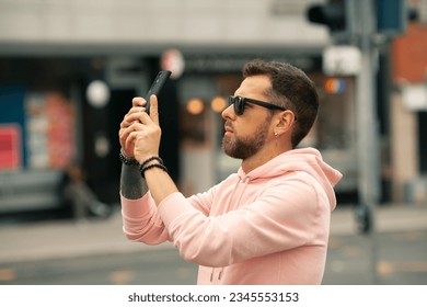 To catch a moment concept. Profile portrait of fashionable young man with beard wearing sunglasses, pink sweatshirt filming video, shooting photograph on city street. Text space. Outside shot - Powered by Shutterstock