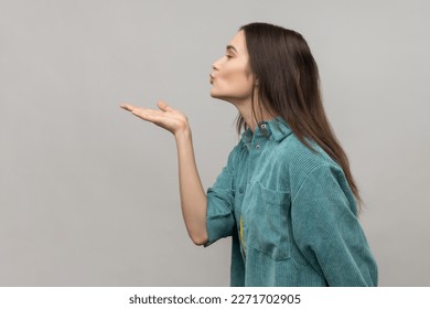 Catch my kiss. Side view of lovely romantic young woman looking ahead air kissing over palms hands, sending love, wearing casual style jacket. Indoor studio shot isolated on gray background.
