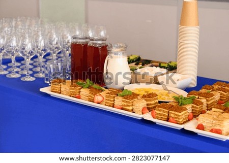 Catering coffee break buffet table with slices of honey cake, milk glass jug, red berry mors drink, empty wine glass rows and paper cup stack.