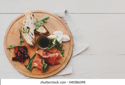 Catering Platter Antipasto With Ham Prosciutto, Bruschetta Bread Toasts And Mozzarella Cheese On Round Wooden Board On White Wood Table Top View With Copy Space. Served Starter Meals, Restaurant Food