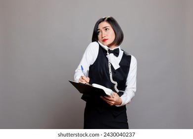 Catering service employee frowning while speaking on landline phone with client and taking notes on clipboard. Waitress looking upwards while chatting with cafe manager on telephone - Powered by Shutterstock