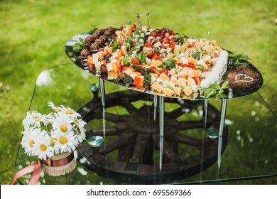 Catering For Wedding Party. Plenty Of Mixed Various Kinds Of Sliced Fruit, Vegetables,  Cheese, Chocolate Sweets On Big Tray On Table Outdoors. Table Decorated With Vase Full Of White Daisies Flowers.