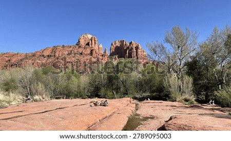 Cathedral Rock rises dramatically against a clear blue sky in Sedona, Arizona, viewed from the red sandstone slickrock along Oak Creek. Visitors relax on the flat rock surface surrounded by spring-green cottonwood trees and desert vegetation. Ideal for travel marketing, tourism campaigns, Southwest lifestyle content, and nature editorial use.
