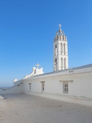 Catholic Christian Church Tower Or Bell Tower Or Belfry Or Steeple Or Campanile In Greek Aegean Island Of Tinos Under Greek Summer White Light With Blue Clear Sky As Background.