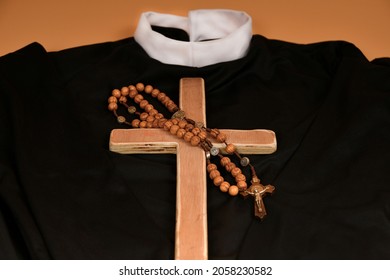 The Catholic Cross And Rosary Lie On The Black Robe Of The Priest On A Beige Background.