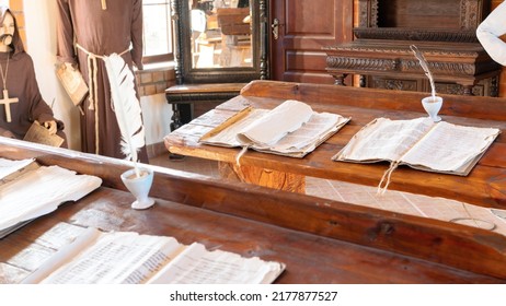 Catholic Monks Are Literacy Teachers In A Medieval School. School In A Catholic Jesuit Monastery. Old Manuscripts And Paper With A Feather Quill Pen Lie On An Aged Wooden Desk In A School Classroom.