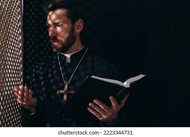 Catholic priest in cassock holding Bible and talking to parishioner in confessional