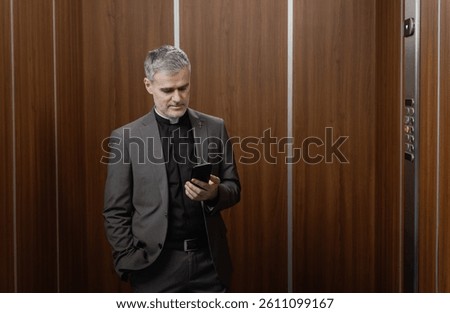 Catholic priest standing in an elevator and using a smartphone, he is checking messages and apps
