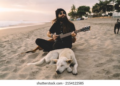 Caucaisan male vegan hippie in trendy accessory performing positive sounds during walk time with mongrel dog at coastline beach, carefree man using folk guitar for playing summer music at seashore