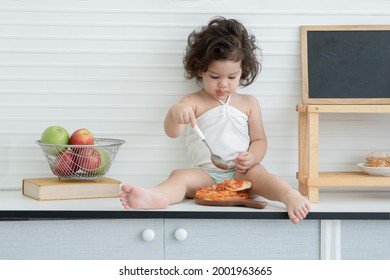 Caucasian Adorable Little Kid Girl Holding Spoon Eating Pizza And Face Mouth Body Mess Up With Ketchup And Sitting On Cupboard At Kitchen At Home. White Background With Black Board, Apples In Basket.