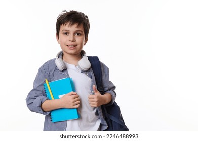 Caucasian adorable primary school student, smart teenage boy in casual wear, with wireless headphones, backpack and school supplies, looks at camera on white background. Copy ad space. Back to school - Shutterstock ID 2246488993