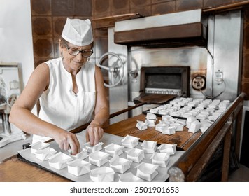 Caucasian adult female baker wearing chef hat and white uniform preparing boxes to fill in a commercial bakery kitchen - Powered by Shutterstock